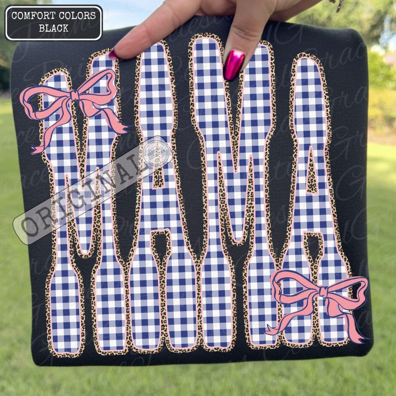 May include: Black t-shirt featuring the word "MAMA" in a blue and white gingham design, edged with leopard print. Pink bows adorn the top left and bottom right. The text "COMFORT COLORS BLACK" is in the top left corner.