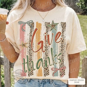 Op de afbeelding: Ivoor Comfort Colors T-shirt met de tekst "Give thanks" in een scriptlettertype. Het ontwerp bevat verticale strepen in roze, oranje en teal, met luipaardprint accenten en ster- en strikdetails. Een grafisch T-shirt met herfstthema.