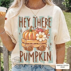 May include: An ivory short-sleeve t-shirt featuring a graphic print of a wooden wagon carrying a pumpkin, an apple, and a white flower. The text "HEY THERE PUMPKIN" is printed in brown above and below the wagon, set against a light blue striped background.