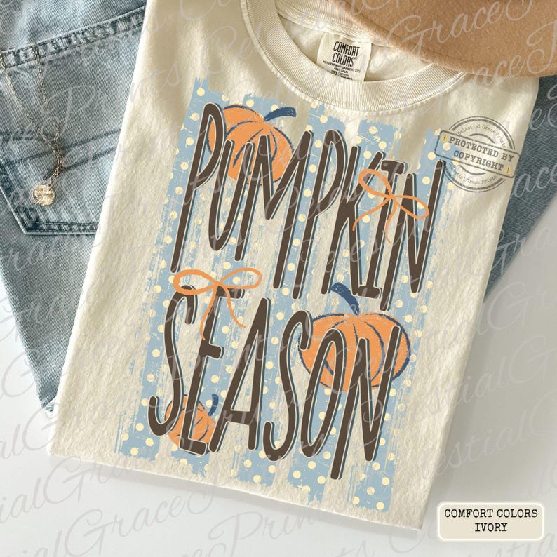 May include: An ivory t-shirt featuring a graphic print with the text PUMPKIN SEASON in dark brown letters. The design includes orange pumpkin illustrations, light blue vertical stripes with white polka dots, and orange line-drawn bows.