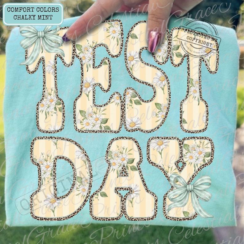 Peut inclure: T-shirt bleu clair avec l'inscription "TEST DAY" dans un motif floral et l&eacute;opard. Les lettres sont remplies de marguerites blanches et de rayures jaunes, soulign&eacute;es d'un imprim&eacute; l&eacute;opard. Le t-shirt a deux n&oelig;uds.
