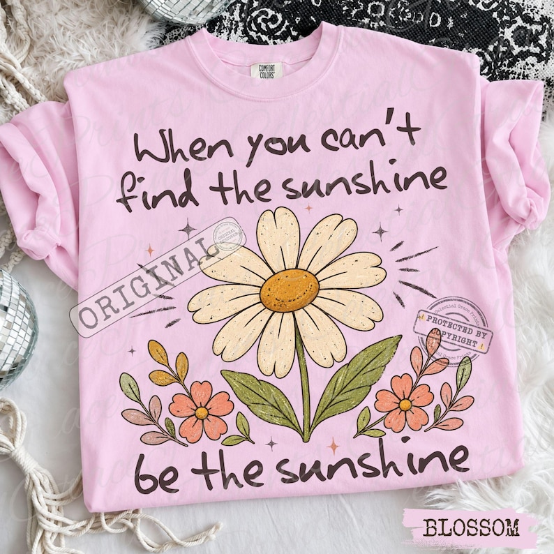 May include: A light pink t-shirt featuring a large daisy illustration in the center with smaller orange flowers at the base. The text reads, When you can't find the sunshine, be the sunshine. The design includes small star accents and green leaves.