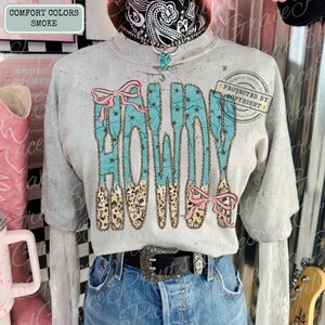 Peut inclure: Un sweat-shirt gris court avec le mot "HOWDY" en turquoise, orn&eacute; de n&oelig;uds roses et d'un motif l&eacute;opard. Le t-shirt est associ&eacute; &agrave; un jean bleu et une ceinture noire avec une boucle argent&eacute;e. Le texte "COMFORT COLORS SMOKE" est dans le coin sup&eacute;rieur gauche.