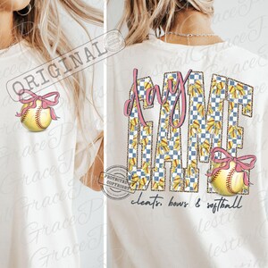 May include: Cream-colored t-shirt with a softball graphic and the words "Day Game." The letters are filled with a blue and white checkered pattern and yellow bows. A softball with a pink bow is on the front and back.
