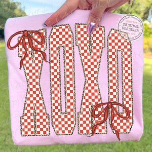May include: A light pink sweatshirt featuring the word "XOXO" in a red and white checkered pattern with a leopard print outline. Red bows are placed on the left and bottom right of the letters. The design is on a light pink background.