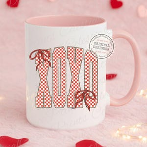 May include: White ceramic mug with a pink interior and handle. The mug features the letters "XOXO" in a red and white checkered pattern, adorned with red bows. The mug has a tag that says "Original Designer".