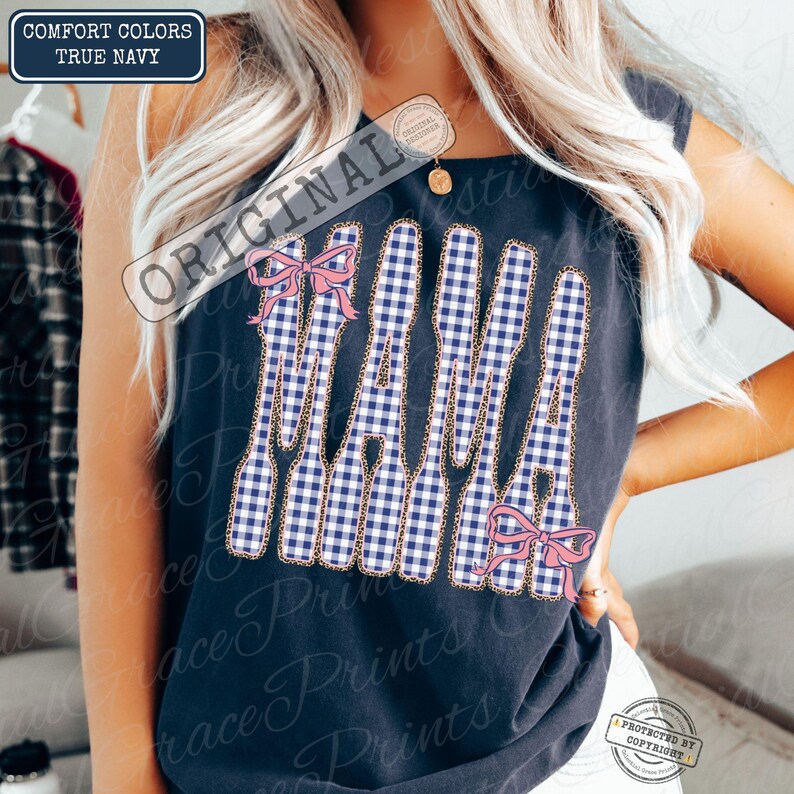 May include: A navy blue vest top featuring the word "MAMA" in a blue and white gingham pattern. The letters are outlined with a gold chain design and decorated with pink bows. The top is labelled "Comfort Colors True Navy."
