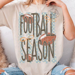 May include: A beige short-sleeve t-shirt featuring a graphic print with the text FOOTBALL SEASON in dark brown letters. The design includes illustrations of three brown footballs, orange line-drawn bows, and a light blue background with yellow dots.
