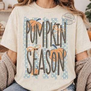 May include: A cream-colored t-shirt featuring a graphic design with the text PUMPKIN SEASON in dark brown letters. The design includes orange pumpkins, thin orange bows, and a background of vertical blue stripes with small yellow polka dots.