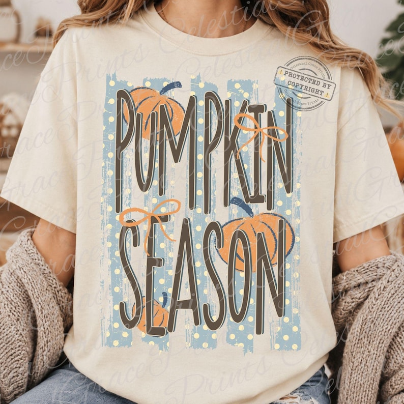 May include: A cream-colored t-shirt featuring a graphic design with the text PUMPKIN SEASON in dark brown letters. The design includes orange pumpkins, thin orange bows, and a background of vertical blue stripes with small yellow polka dots.