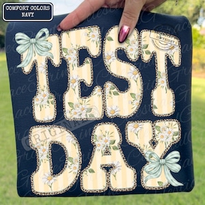 Peut inclure: T-shirt bleu marine avec l'inscription "TEST DAY" dans un motif floral et l&eacute;opard. Les lettres jaunes sont orn&eacute;es de rayures verticales et de fleurs de marguerites blanches. Le t-shirt est orn&eacute; de n&oelig;uds bleu clair.
