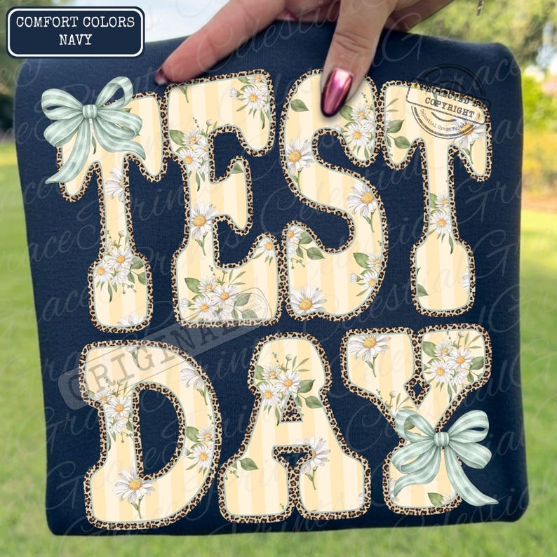 Peut inclure: T-shirt bleu marine avec l'inscription "TEST DAY" dans un motif floral et l&eacute;opard. Les lettres jaunes sont orn&eacute;es de rayures verticales et de fleurs de marguerites blanches. Le t-shirt est orn&eacute; de n&oelig;uds bleu clair.