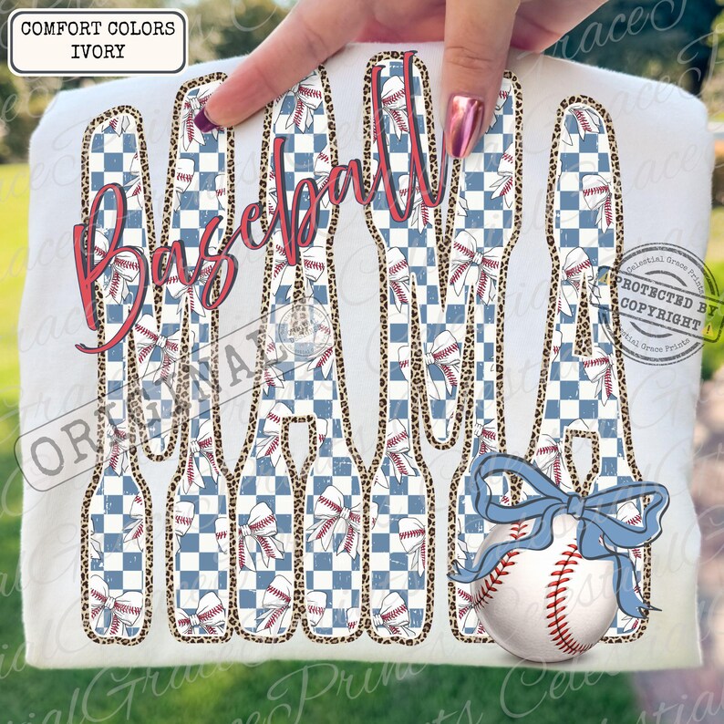May include: Ivory-colored t-shirt featuring the word "Baseball" in a cursive, red font. The letters are filled with a blue and white checkered pattern and baseball-themed designs. A baseball with a blue bow is also visible.