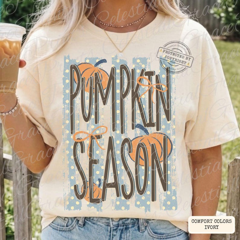 May include: An ivory short-sleeve t-shirt featuring a graphic print with the text PUMPKIN SEASON in dark brown letters. The design includes light blue vertical stripes with white polka dots, orange pumpkin illustrations, and thin orange bows.