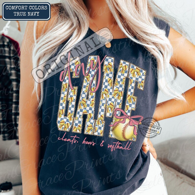 May include: Navy blue tank top with the words "Game Day" in pink script and a checkered pattern. A softball with a pink bow is featured below the text. The words "cleats, bows & softball" are written in pink script.