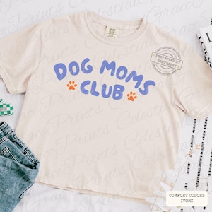 May include: Cream-colored t-shirt with the text "DOG MOMS CLUB" in blue, featuring orange paw prints. The shirt is a Comfort Colors brand in ivory. A casual, everyday wear item for dog lovers.