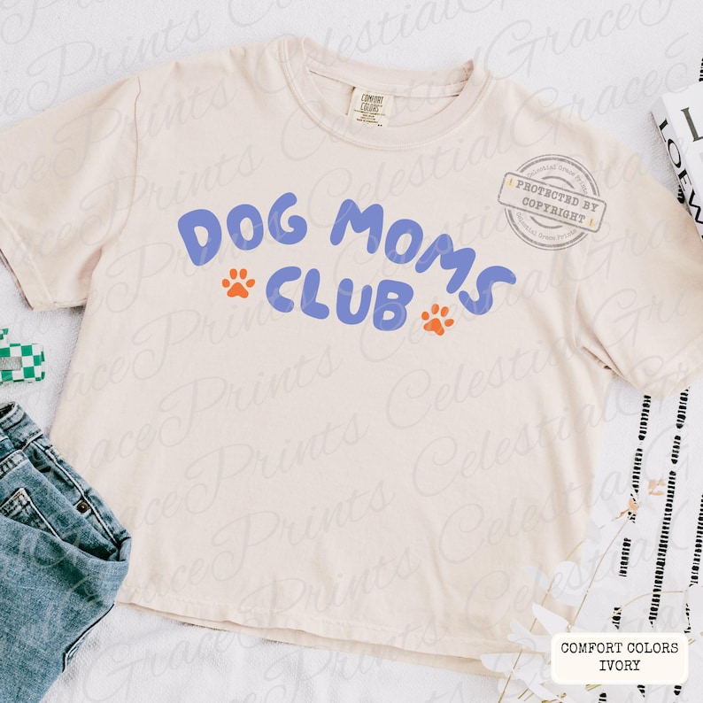 May include: Cream-colored t-shirt with the text "DOG MOMS CLUB" in blue, featuring orange paw prints. The shirt is a Comfort Colors brand in ivory. A casual, everyday wear item for dog lovers.
