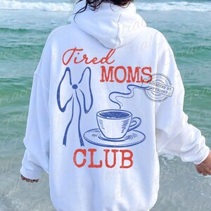 May include: White hoodie with the words "Tired Moms Club" in red and blue. The design includes a blue bow, a steaming coffee cup, and a saucer. The hoodie is worn by a person standing near the ocean.