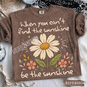 May include: A brown short-sleeve t-shirt featuring a large white daisy graphic with a yellow center, surrounded by smaller pink flowers and green leaves. The text reads, "When you can't find the sunshine, be the sunshine," in a cream-colored font.