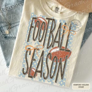 May include: An ivory t-shirt featuring a graphic with the text FOOTBALL SEASON in dark brown letters. The design includes three brown football illustrations, a light blue background with white polka dots, and two orange line-drawn bows.