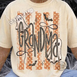 May include: A cream-colored t-shirt featuring a graphic print with the word GRANDMA in large dark letters. The design includes vertical orange stripes with white polka dots, two white ghost illustrations, two black bats, and two black bows.