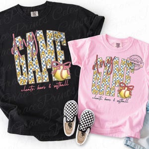 May include: Two t-shirts, one black and one pink, with a softball theme. The shirts feature the words "Game Day" in a checkered pattern, a softball with a pink bow, and the text "cleats, bows & softball". Checkerboard slip-on shoes are in front.