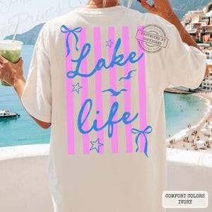 Puede incluir: Camiseta de color marfil con un dise&ntilde;o rosa y azul. La parte posterior de la camiseta presenta rayas verticales rosas, texto azul "Lake Life", estrellas, p&aacute;jaros y lazos. La camiseta est&aacute; hecha de material Comfort Colors.