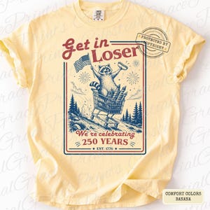 May include: Banana-colored t-shirt with a graphic featuring a raccoon in a shopping cart, holding an American flag and a hot dog. The text reads "Get in Loser" and "We're celebrating 250 years." The shirt is a Comfort Colors brand.