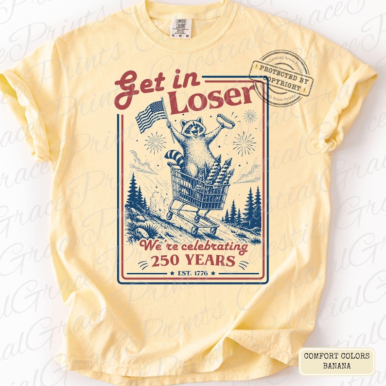 May include: Banana-colored t-shirt with a graphic featuring a raccoon in a shopping cart, holding an American flag and a hot dog. The text reads "Get in Loser" and "We're celebrating 250 years." The shirt is a Comfort Colors brand.