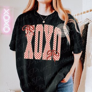 May include: Black t-shirt with the word "XOXO" in a red and white checkered pattern. The letters are outlined in a gold color and have red bow accents. A neon sign that says "OXOX" is in the background.