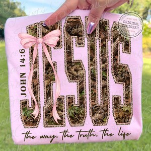 May include: A light pink sweatshirt featuring the word "JESUS" in a camouflage pattern with leopard print trim. The text "JOHN 14:6" and "the way, the truth, the life" are also visible. A pink bow is on the left side.