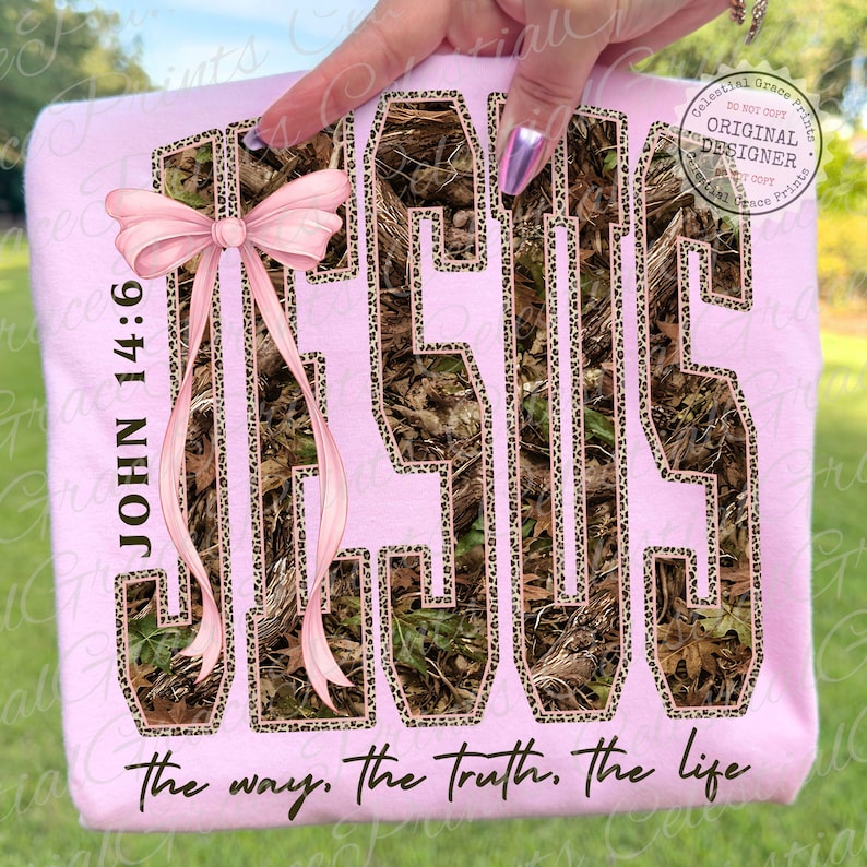 May include: A light pink sweatshirt featuring the word "JESUS" in a camouflage pattern with leopard print trim. The text "JOHN 14:6" and "the way, the truth, the life" are also visible. A pink bow is on the left side.