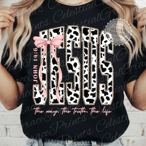 Puede incluir: Camiseta negra con la palabra "JESUS" en un dise&ntilde;o de estampado de vaca, acentuada con un lazo rosa. El texto "JOHN 14:6" est&aacute; a la izquierda, y "the way, the truth, the life" est&aacute; en la parte inferior.