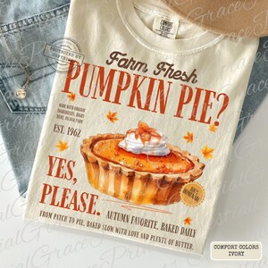 May include: An ivory t-shirt featuring a graphic of a pumpkin pie with whipped cream, surrounded by orange maple leaves. Text reads: Farm Fresh PUMPKIN PIE? EST. 1962 YES, PLEASE. MADE WITH ORGANIC INGREDIENTS, RIGHT HERE, ON OUR FARM. AUTUMN FAVORITE, BAKED DAILY.