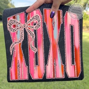 May include: Black t-shirt with the word "MAMA" in large, colorful letters. The letters are vertically striped in shades of pink, orange, and blue, with a leopard print bow. The design is outlined in leopard print.