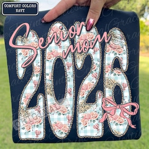 Può includere: Felpa blu navy con la scritta "Senior Mom 2026" in un design rosa, floreale e leopardato. Il design include un fiocco rosa e palloncini a forma di cuore. L'angolo in alto a sinistra ha un rettangolo blu navy con "Comfort Colors Navy".
