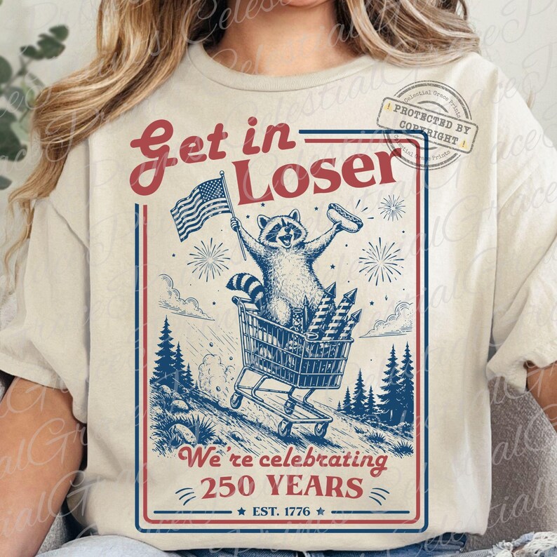 May include: Beige t-shirt with a graphic featuring a raccoon in a shopping cart, waving an American flag and holding a hot dog. The text reads "Get in Loser" and "We're celebrating 250 YEARS".