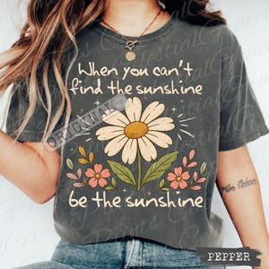 May include: A charcoal gray short-sleeve t-shirt featuring a large daisy graphic in the center. The text reads, When you can't find the sunshine be the sunshine. The design includes small pink flowers, green leaves, and star accents in a light cream color.
