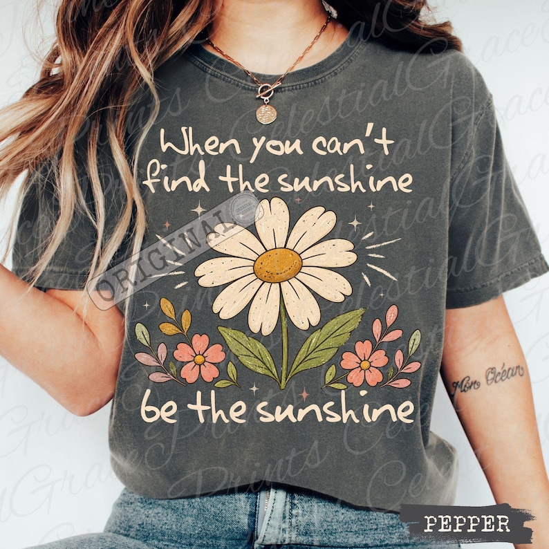 May include: A charcoal gray short-sleeve t-shirt featuring a large daisy graphic in the center. The text reads, When you can't find the sunshine be the sunshine. The design includes small pink flowers, green leaves, and star accents in a light cream color.