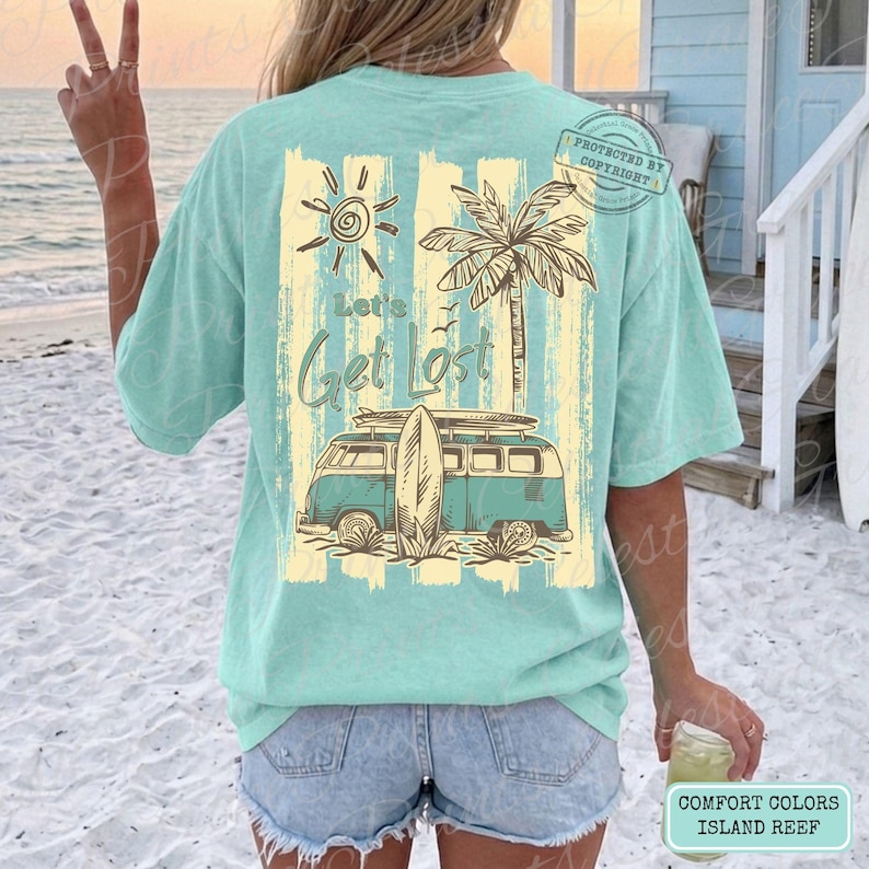 May include: A light teal t-shirt with a graphic featuring a sun, palm tree, and a vintage van with a surfboard. The text "Let's Get Lost" is printed on the shirt. The shirt is a Comfort Colors Island Reef.