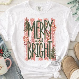 May include: A white short-sleeve t-shirt featuring a graphic print with the text MERRY and BRIGHT in dark green letters. The design includes red and white striped candy canes, green Christmas trees, and vertical red polka dot stripes.