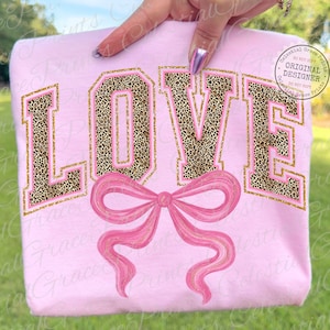 May include: A light pink sweatshirt with the word "LOVE" in large, block letters. The letters have a leopard print pattern with gold and pink trim. A pink bow is below the word. The image is held by a hand.