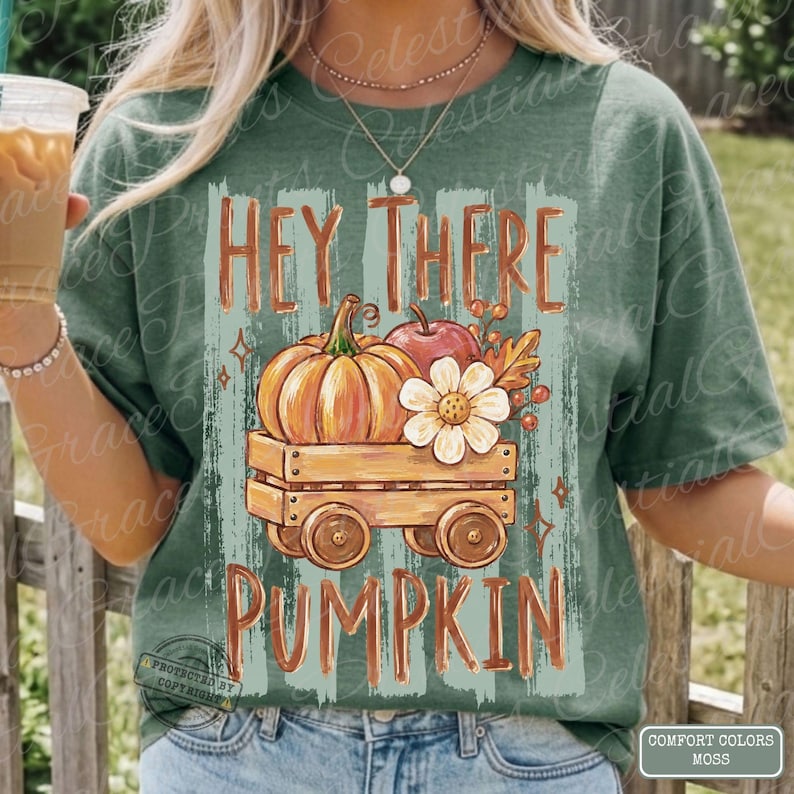 May include: A sage green t-shirt featuring a graphic of a wooden wagon carrying a large orange pumpkin, a red apple, and a white flower. The text HEY THERE PUMPKIN is printed in orange above and below the wagon illustration on a light green background.