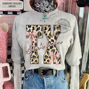 May include: A gray t-shirt with the text "TX She's from" in a pink script. The letters TX are filled with a leopard print design and adorned with pink bows. The shirt is paired with blue jeans and a black belt.