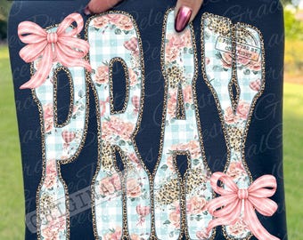 Puede incluir: Una camiseta azul marino con la palabra PRAY en letras grandes. Las letras tienen un patrón de cuadros vichy azul claro y flores rosas con un borde de estampado de leopardo. Un lazo a rayas rosa está unido a la P y a la Y.