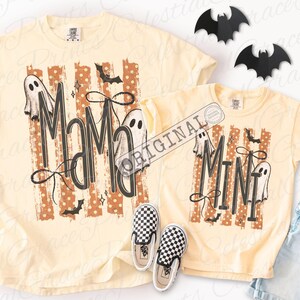 May include: Two pale yellow t-shirts featuring a graphic print with orange polka-dot stripes, black bats, and white ghosts. The larger shirt displays the text MAMA in black, while the smaller shirt displays the text MINI in black. Both shirts include black bow accents.