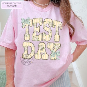 Peut inclure: T-shirt rose clair avec l'inscription "TEST DAY" dans un motif jaune et floral. Les lettres ont un motif ray&eacute; et sont d&eacute;cor&eacute;es de fleurs blanches et de n&oelig;uds. Le t-shirt est &eacute;tiquet&eacute; "COMFORT COLORS BLOSSOM".