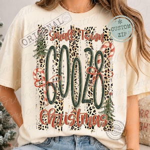 Puede incluir: Una camiseta color crema con un gráfico que incluye el texto Small Town 60028 Christmas. El diseño presenta rayas verticales con estampado de leopardo, dos árboles de Navidad verdes y dos bastones de caramelo, con el número 60028 en verde oscuro.