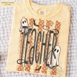Puede incluir: Una camiseta de color amarillo mantequilla con un estampado gr&aacute;fico que incluye la palabra TEACHER en letras negras grandes. El dise&ntilde;o presenta dos fantasmas blancos, rayas verticales naranjas con puntos blancos y murci&eacute;lagos.
