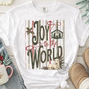Puede incluir: Una camiseta blanca de manga corta con un estampado gr&aacute;fico que incluye el texto Joy to the World en verde oscuro y rojo. El dise&ntilde;o presenta una silueta de bel&eacute;n, un peque&ntilde;o cordero, hojas de acebo y rayas verticales en tonos apagados.